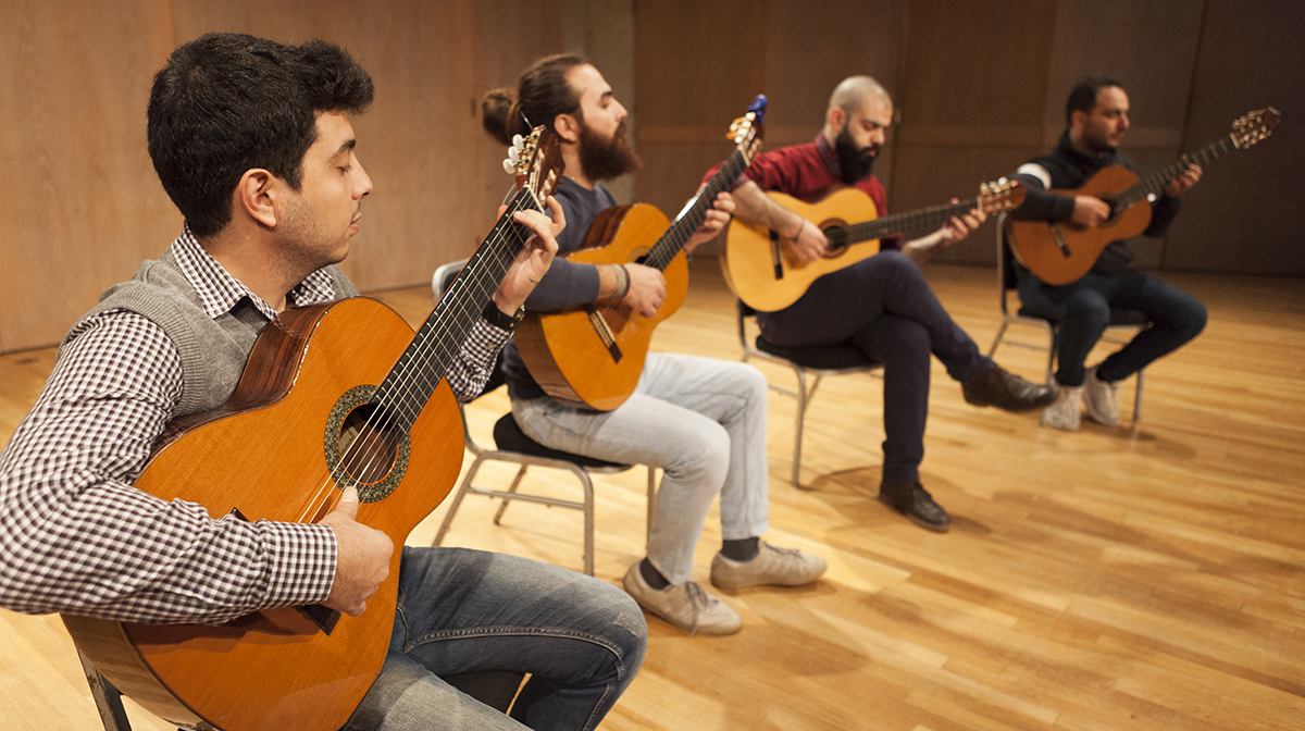 The four male guitarists in Orentes sit on chairs, playing the guitar on a stage with wooden floors.